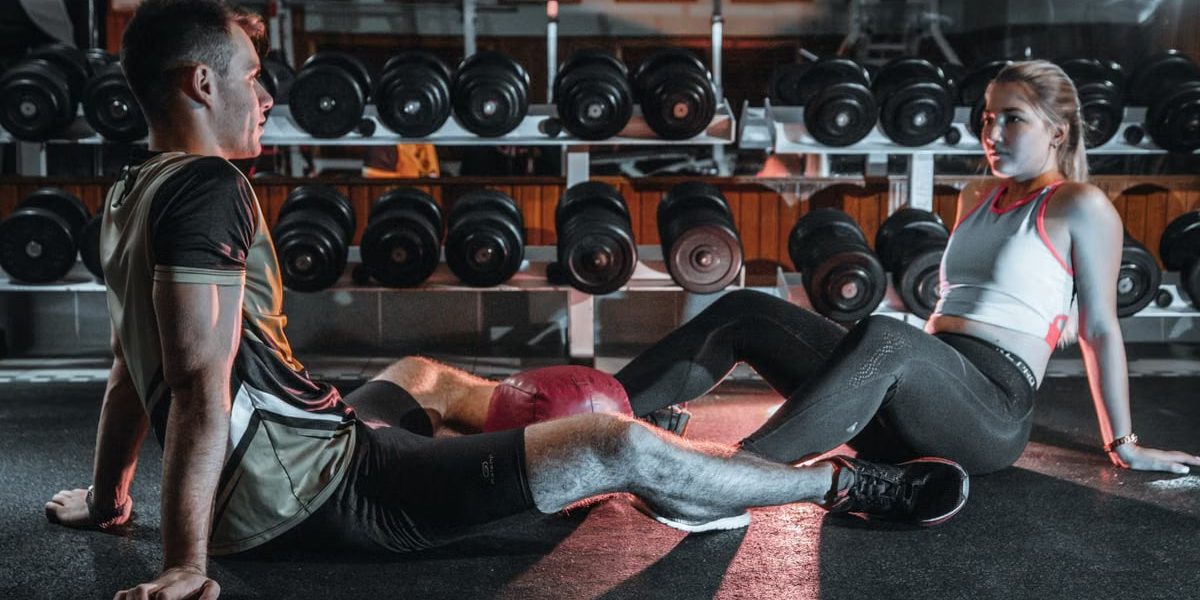 A man and woman resting together on the gym floor, surrounded by dumbbells.