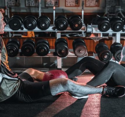 A man and woman resting together on the gym floor, surrounded by dumbbells.